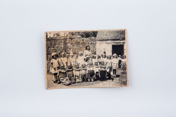 Grup de copii la serbare împreună cu învățătoarea lor, în curtea școlii. Băieții au coifuri pe cap. Fotografie de exteri